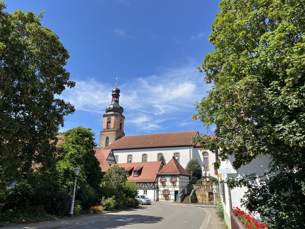 Das Bild zeigt eine Sommerliche Ansicht der Hauptstraße von Rheinzabern. Der Blick geht über die Erlenbachbrücke in Richtung der Kirche. Vor der kirche schmiegen sich Fachwerkhäuser an die Straße. darüber trohnt die Kirche mit ihrem geschwungenem Turmhelm.