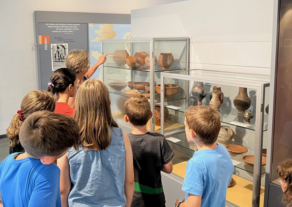 Die Kursleiterin Regina Antoni zeigt den Kindern der Römerwoche die verschiedenen Oberflächentexturen römischer Keramik. Sie stehen dazu im Museum vor einer Vitrine und sehen sich antike Gefäße an.
