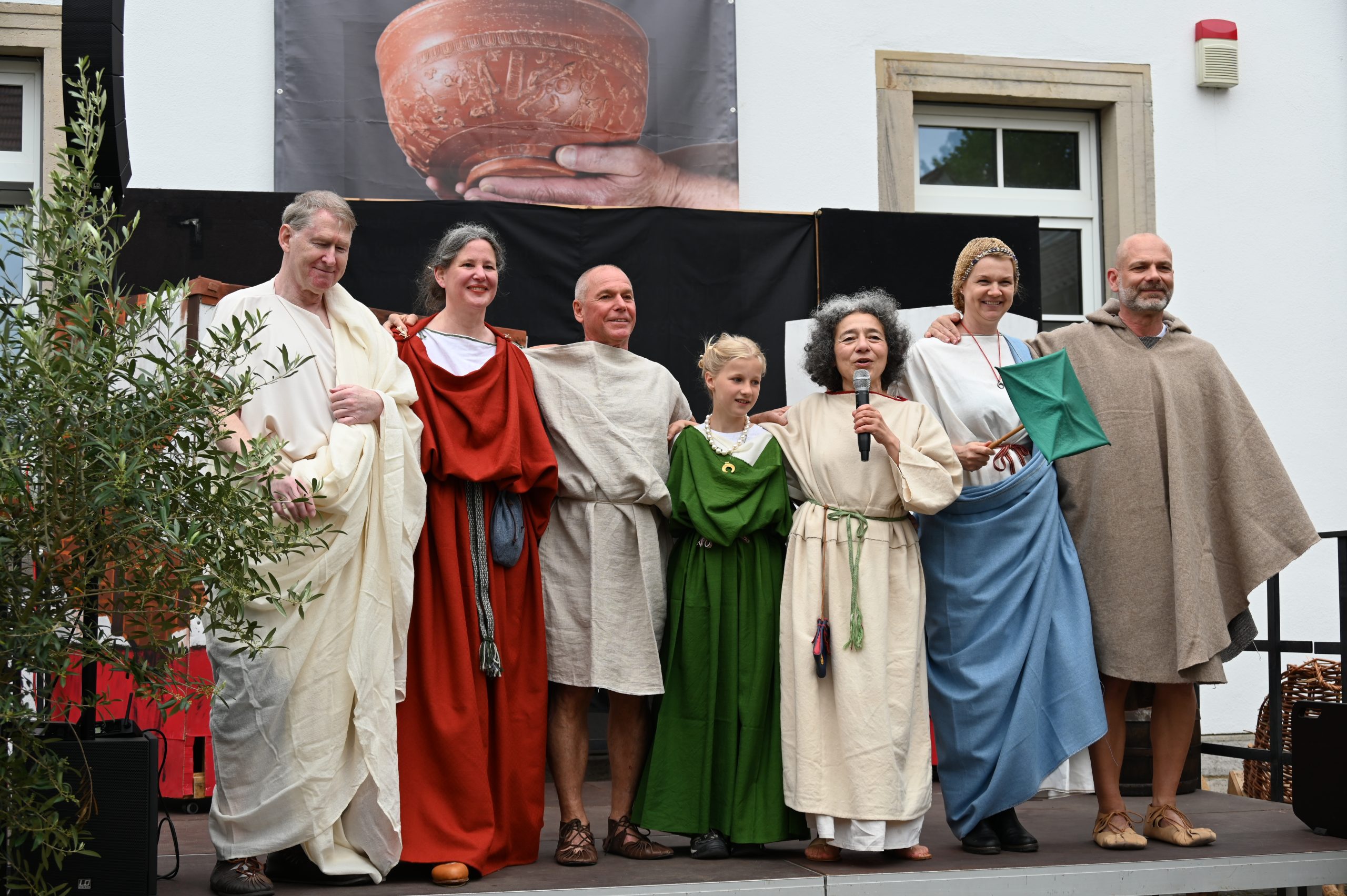 Zu sehen sind die Teilnehmer der römischen Modenschau: Ein Mann in Toga, eine Frau in provinzialrömischer Kleidung, ein Mann in römischer Arbeitskleidung, ein Mädchen in provinzialrömischer Kleidung, die Moderatorin Gisela Michel, eine Frau in Stadtrömischer Kleidung und ein Mann in provinzialrömischer Kleidung.