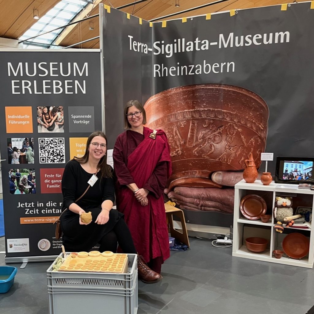 Der Messestand des Museums war mit großen Plakaten und großformatigen Bilern der römischen Terra-Sigillata-Schüsseln dekoriert. Zwei Frauen lachen in die Kamera. Eine davon ist mit einem roten Gewand römisch gekleidet.